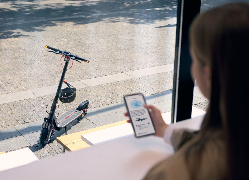 Hulajnoga elektryczna SEGWAY Ninebot F3 Pro D 70km 1200W Amortyzacja 10 cali Czarny miasto osoba siedzi w budynku trzyma telefon w dłoni przed budynkiem stoi hulajnoga wbudowany moduł Bluetooth aplikacja mobilna Segway-Ninebot Smart App na systemy Android i iOS