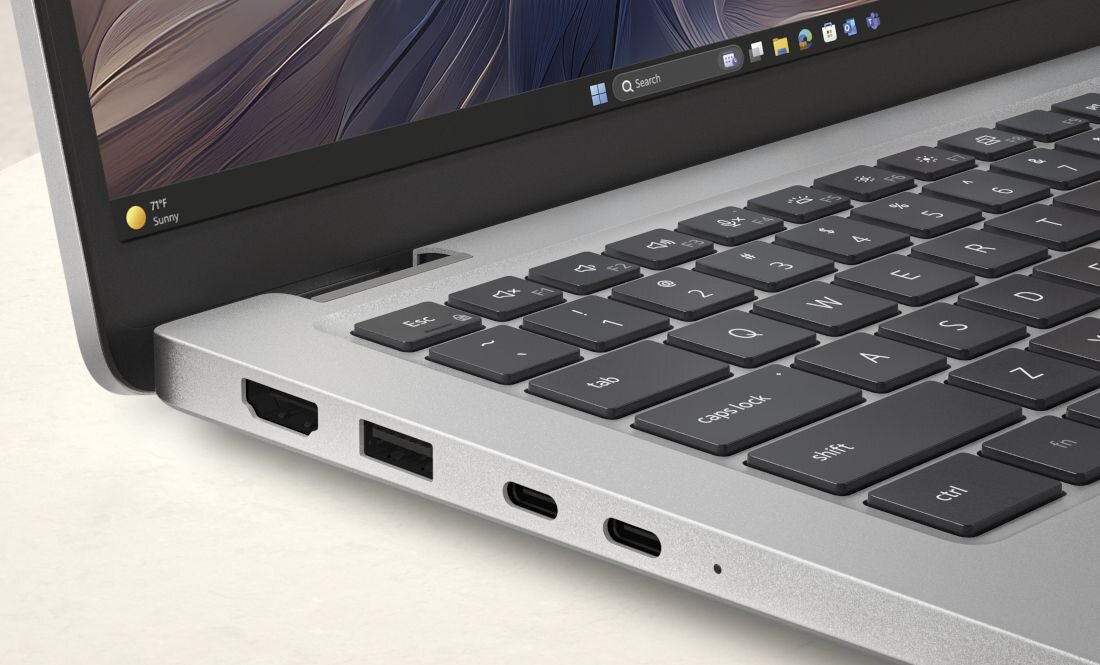 Na obrazie widoczny jest srebrny laptop z klawiaturą i kilkoma portami po boku. Na ekranie widać pasek z ikonami systemu Windows 11.