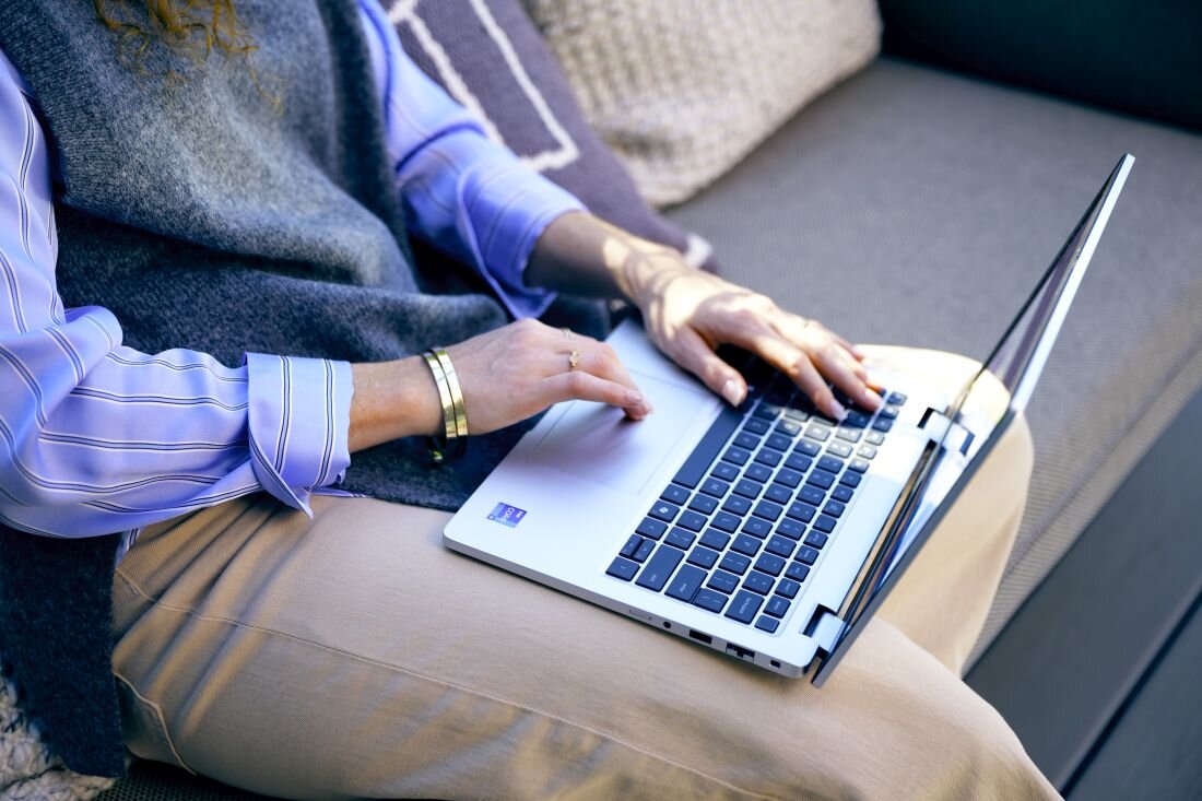 Osoba siedzi na kanapie i używa laptopa z klawiaturą w kolorze czarnym i srebrną obudową. Na laptopie widoczne jest logo producenta w formie naklejki.