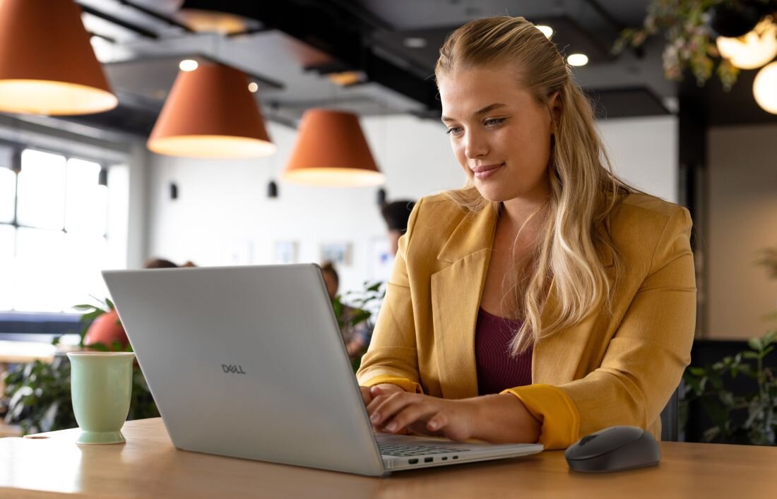 Osoba pracuje na laptopie marki Dell w jasnym wnętrzu z wiszącymi lampami. Obok laptopa znajduje się myszka i zielony kubek.