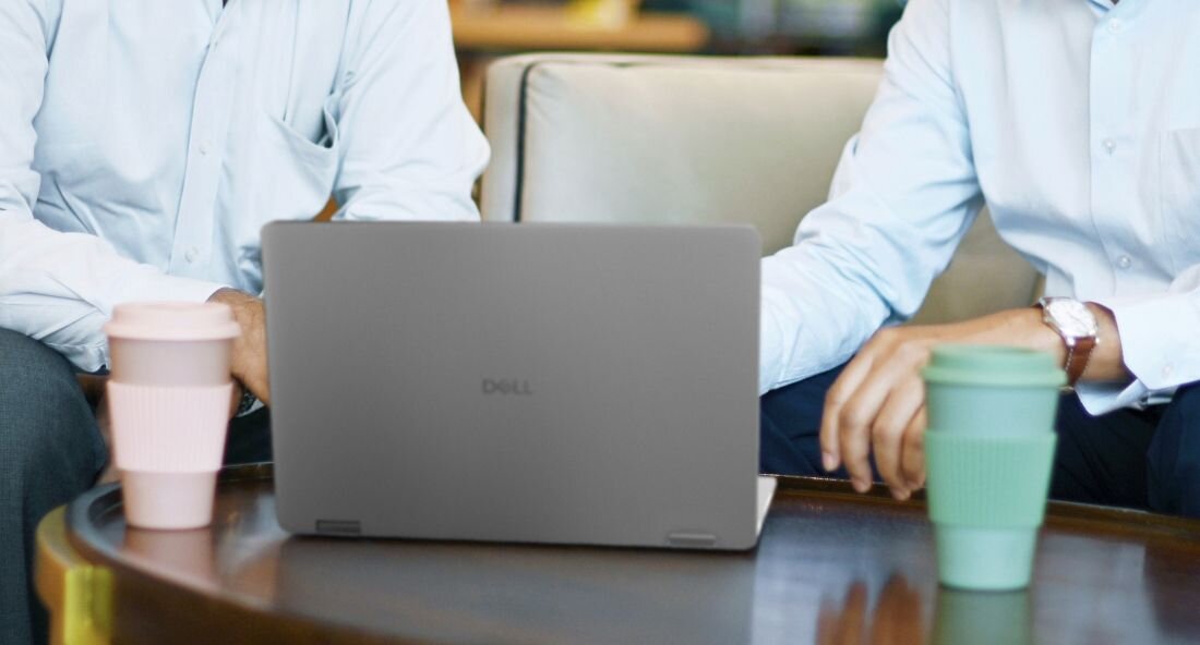 Na stole stoi otwarty laptop z widocznym logo \'DELL\'. Obok znajdują się dwa kubki z pokrywkami, a w tle widać dwie osoby w koszulach.