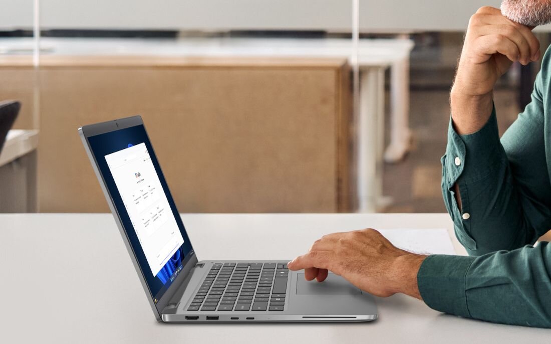 Osoba korzysta z laptopa na biurku, na ekranie widoczny jest interfejs systemu operacyjnego. W tle znajduje się rozmyte wnętrze biura.