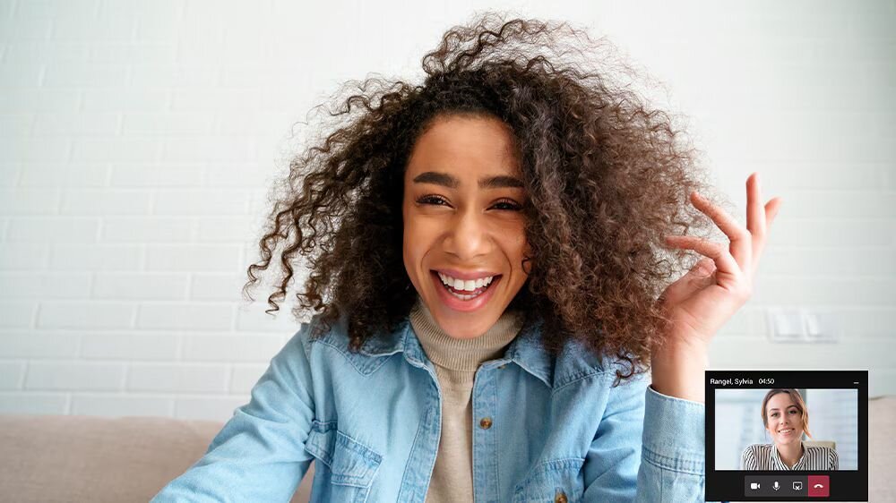 Osoba z kręconymi włosami uśmiecha się do kamery, siedząc przed białą ceglaną ścianą. W prawym dolnym rogu ekranu widoczny jest mały podgląd wideorozmowy z inną osobą.