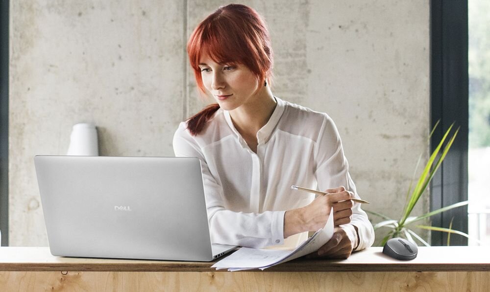 Kobieta pracuje na laptopie DELL Pro 14 PC14250 zapisując notatki w industrialnym biurze – DELL Trusted Workspace zabezpieczenia BIOS ochrona łańcucha dostaw