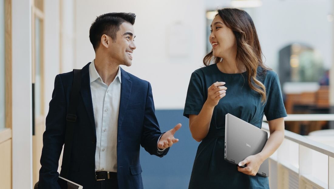 Dwie osoby idą korytarzem, jedna z nich trzyma laptopa z widocznym logo DELL. Obie osoby są uśmiechnięte i rozmawiają ze sobą.