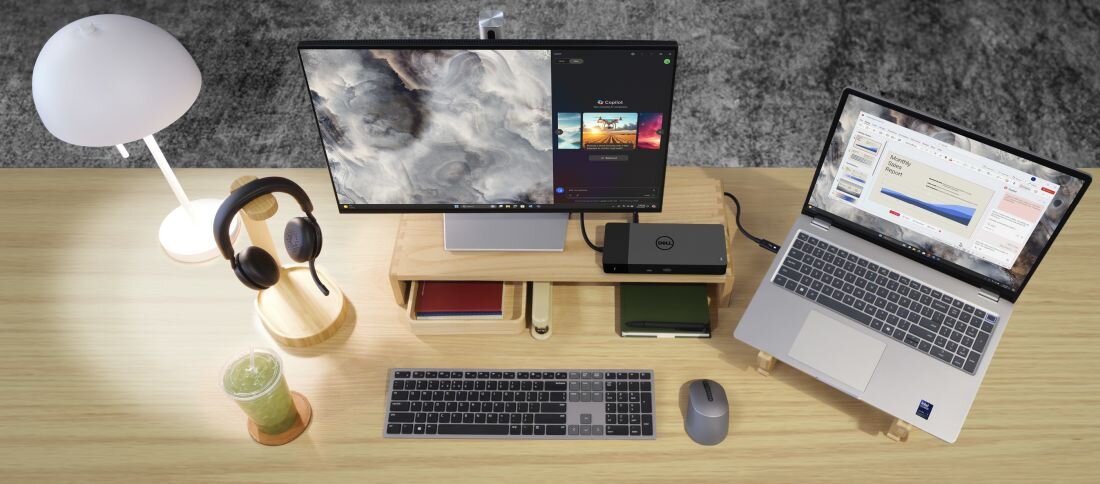 Na biurku znajduje się laptop z otwartym ekranem, obok którego stoi monitor z wyświetlonymi grafikami. Obok monitora leżą słuchawki, klawiatura, myszka oraz szklanka z zielonym napojem.