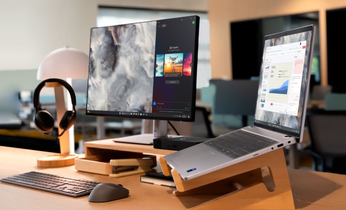 Na biurku znajduje się laptop na drewnianej podstawce, obok monitora z wyświetlonymi aplikacjami. W tle widoczna jest lampa, klawiatura, myszka i słuchawki.