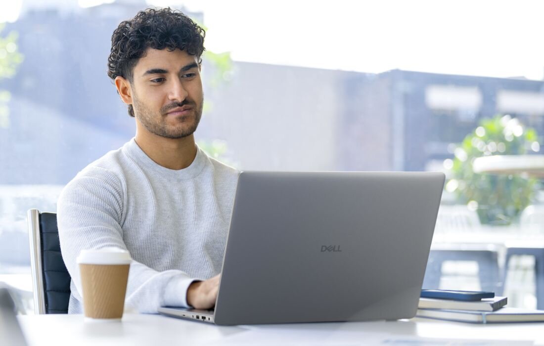 Osoba siedzi przy biurku, używając laptopa z widocznym logo \'Dell\'. Obok laptopa stoi kubek z napojem i leżą notatniki.