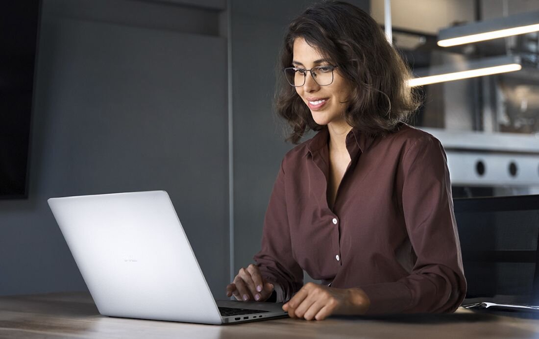 Osoba siedzi przy biurku i korzysta z laptopa o srebrnej obudowie. W tle widoczne są elementy wnętrza biurowego.