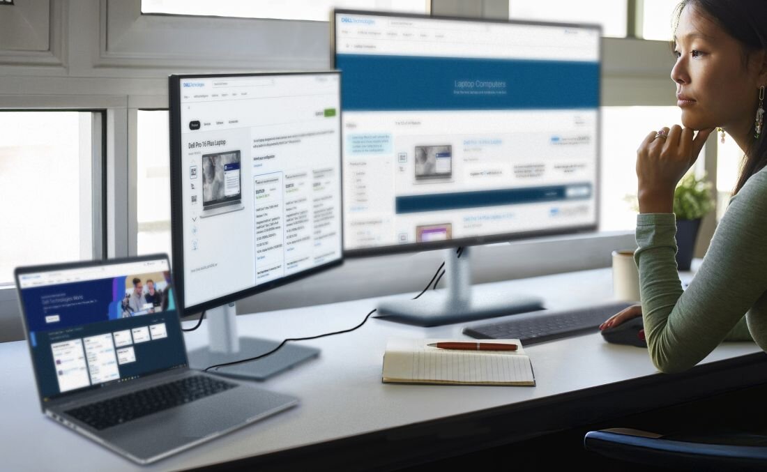 Na biurku znajduje się otwarty laptop oraz dwa duże monitory, na których wyświetlane są strony internetowe. Obok laptopa leży notatnik i długopis, a w tle widać osobę siedzącą przy biurku.