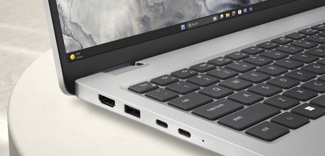 Zbliżenie na klawiaturę i porty boczne srebrnego laptopa. Na ekranie widoczny jest pasek z ikonami systemu Windows.