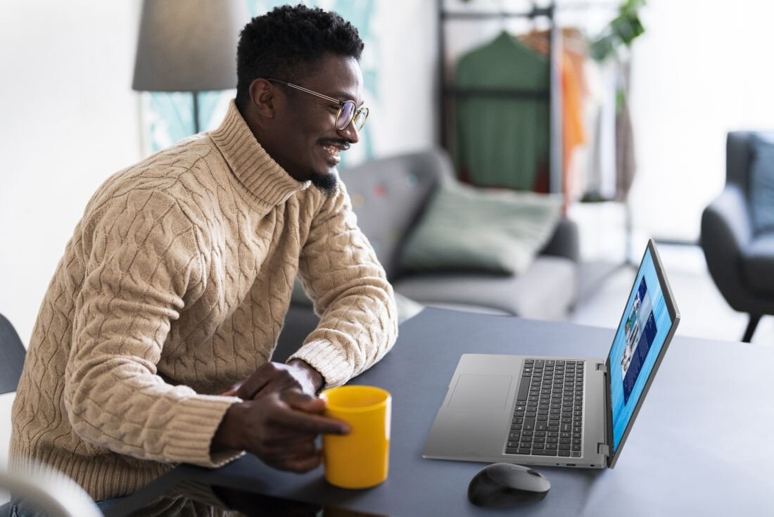 Osoba siedzi przy stole z otwartym laptopem i trzyma żółty kubek. W tle widać rozmyte wnętrze z sofą i wieszakami na ubrania.