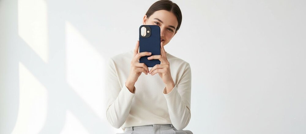 Mężczyzna w białym swetrze i jasnych spodniach trzyma przed twarzą telefon w granatowym etui