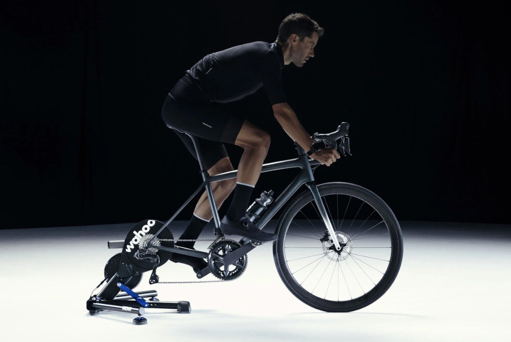 Trenażer rowerowy WAHOO Kickr Smart Trainer V6 WFBKTR122 ciemne tło osoba jedzie na rowerze rower umieszczony na trenażerze realistyczny trening trening rowerowy w domy wszechstronność wygoda bezpieczeństwo