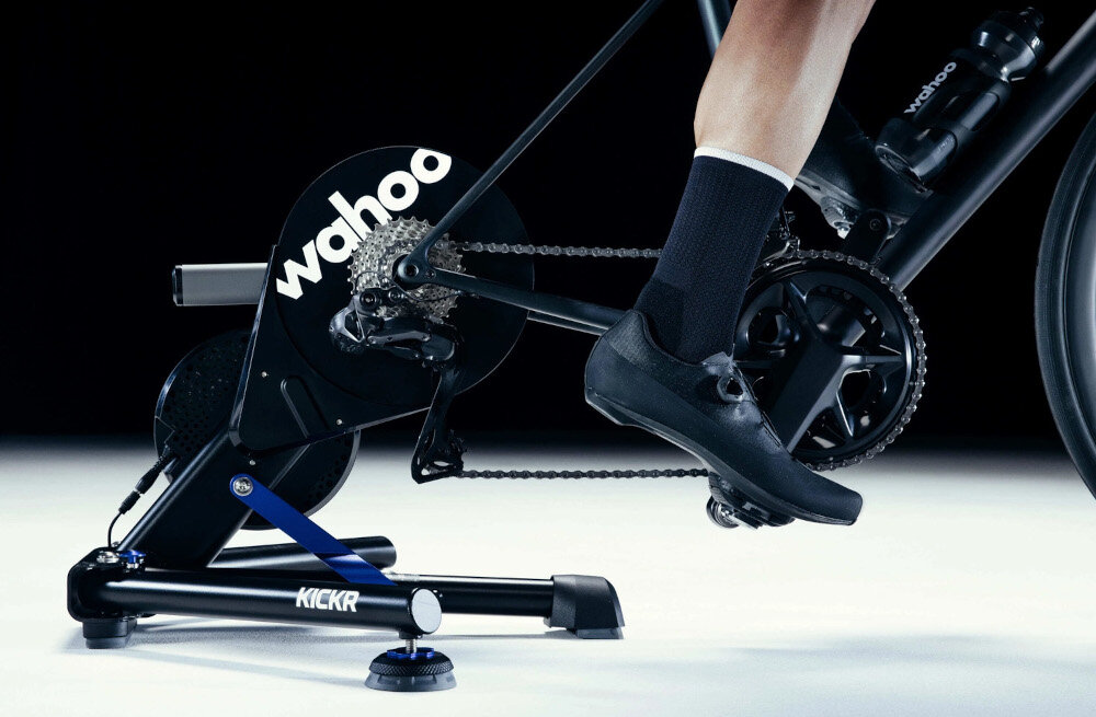 Trenażer rowerowy WAHOO Kickr Smart Trainer V6 WFBKTR122 ciemne tło osoba jedzie na rowerze rower umieszczony na trenażerze trenażer rowerowy rolkowy tylne koło spoczywa na rolkach przednie w uchwycie stabilizującym odwzorowuje naturalne odczucia jazdy realistyczny trening w domu