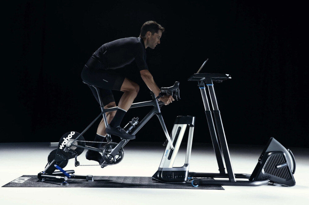 Trenażer rowerowy WAHOO Kickr Smart Trainer V6 WFBKTR122 ciemne tło osoba jedzie na rowerze rower umieszczony na trenażerze współpracuje z osiami 12x142, 12x148 i standardowymi szybkozamykaczami 130/135 mm uniwersalność sprzętu intuicyjna aplikacja Wahoo Fitness