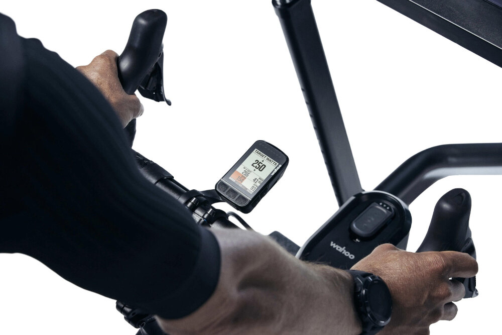 Trenażer rowerowy WAHOO Kickr Smart Trainer V6 WFBKTR122 białe tło osoba jedzie na rowerze rower umieszczony na trenażerze na trenażerze licznik wspópracuje z symulatorem KICKR CLIMB wentylatorem KICKR HEADWIND i licznikami ELEMNT aplikacja Wahoo SYSTM personalizowane treningi z elementami siłowymi mentalnymi i jogą
