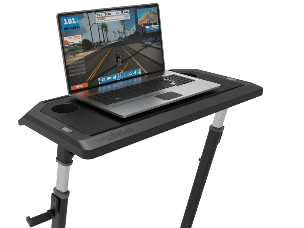 Biurko do trenażera WAHOO KickR Desk V2 WFDESK2 zbliżenie na blat na blacie stoi laptop widoczny uchwyt na kable dwa wieszaki na koła dwa miejsca na butelki uchwyty na kable USB porządek wokół trenażera akcesoria są w zasięgu ręki podstawa biurka o wymiarach 69,8 × 74,9 cm maksymalny udźwig do 22,67 kg