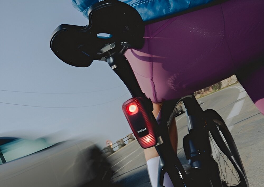 Wsteczny radar rowerowy WAHOO TrackR Radar Smart WFTRACKRRDR1 miasto droga ulica osoba jedzie na rowerze zbliżenie na radar umieszczony pod sztycą siodełka fale radiowe do wykrywania nadjeżdżających pojazdów z odległości nawet do 150 metrów planować manewry i utrzymać bezpieczną linię jazdy użytkownik reaguje szybciej radar automatycznie uruchamia sekwencję błysków ostrzegawczych poprawia widoczność