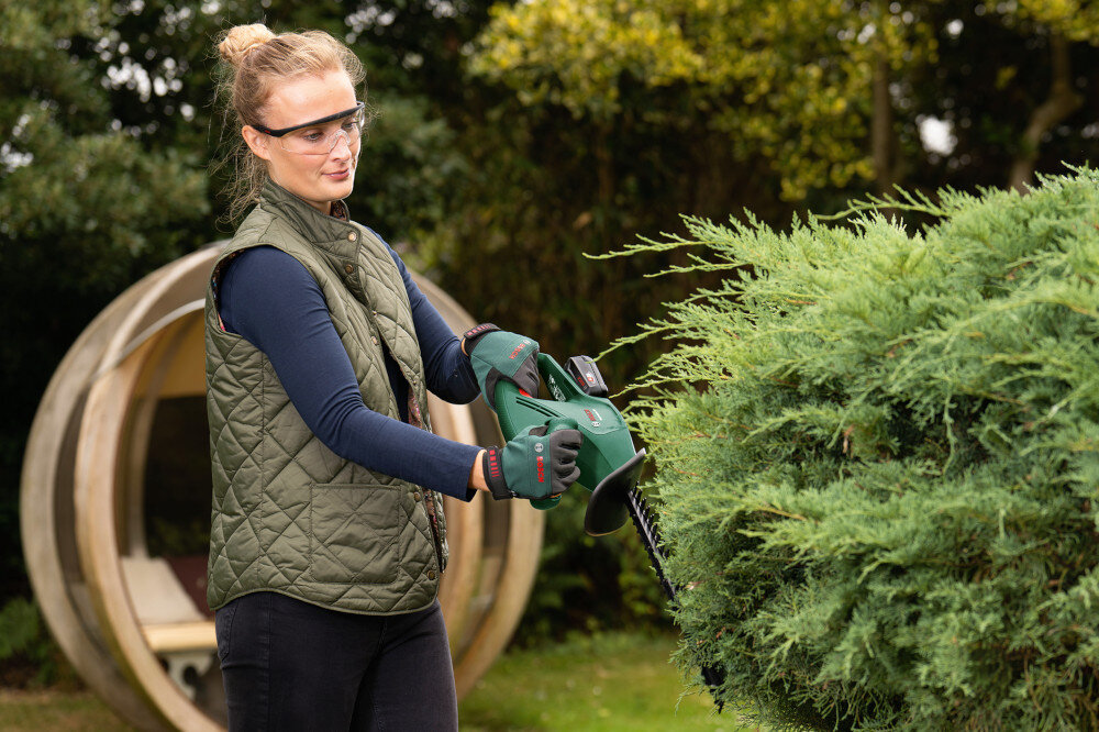 Nożyce do żywopłotu BOSCH EasyHedgeCut 18V-44-11 ogród osoba tnie żywopłot trzymając nożyce w dłoniach w tle drzewa silnik szczotkowy stabilna i równomierna praca łatwy w obsłudze i serwisowaniu wystarczająca moc do standardowych zadań pielęgnacyjnych płynne cięcie i skuteczne formowanie roślin