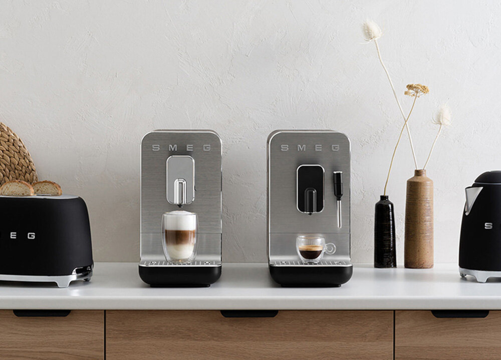 Na blacie kuchennym znajdują się dwa ekspresy do kawy SMEG, obok czajnika i tostera tej samej marki. Ekspresy mają srebrne fronty, a na jednym z nich stoi filiżanka z kawą, na drugim szklanka z latte.
