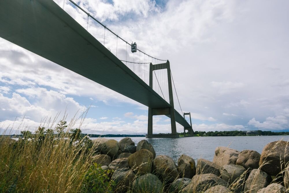 Zdjęcie przedstawia most wiszący nad wodą, z widocznymi pylonami i linami nośnymi. Na pierwszym planie znajdują się kamienie i trawa, a w tle widać chmury na niebie.