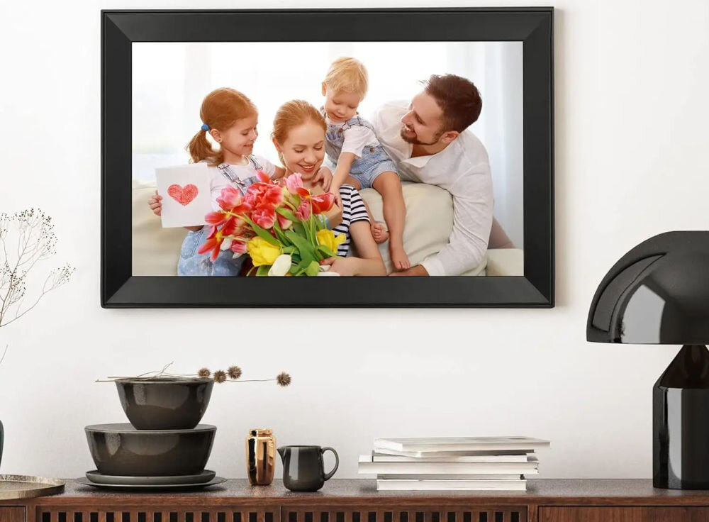 Ramka cyfrowa BLOW 15.6 Frameo Wi-Fi prezentacja ramki ze zdjęciem powieszonej na jasnej ścianie pod ramką półka RTV z rozłożonymi rzeczami aplikacja FRAMEO przesyłanie zdjęć do ramki przez Wi-Fi reakcje pod zdjęciami osobisty charakter