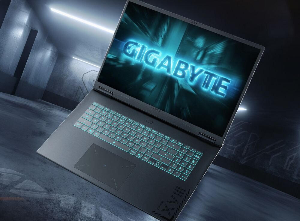 Laptop z podświetlaną klawiaturą i ekranem wyświetlającym logo \'GIGABYTE\'. Otoczenie przypomina nowoczesne, industrialne wnętrze.