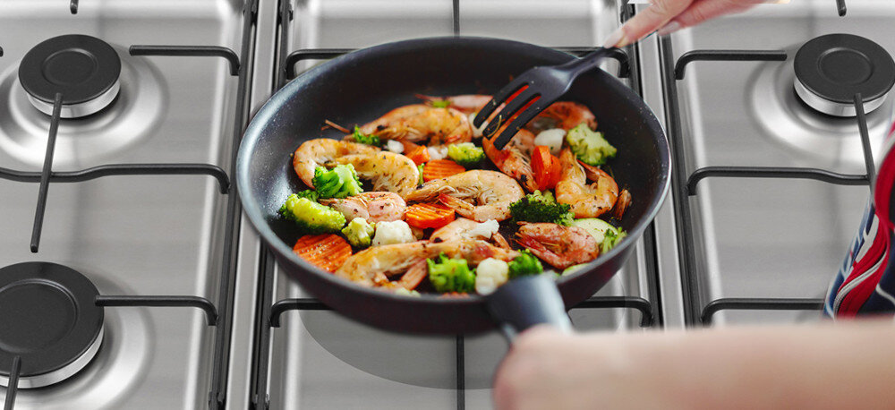 KUCHNIA DAEWOO F96CGDTE19INM-EE Zdjęcie przedstawia zbliżenie na patelnię ustawioną na palniku gazowym, na której smażone są krewetki z mieszanką warzyw, takich jak brokuły, marchewka i kalafior. Zdjęcie przedstawia także fragment płyty gazowej z kilkoma palnikami oraz dłonie osoby mieszającej potrawę plastikową łopatką płyta gazowa 5 palników palnik wok regulacja płomienia ruszty emaliowane