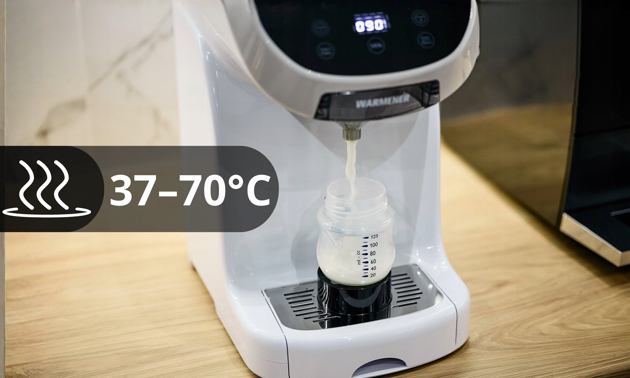 Na blacie stoi ekspres do mleka modyfikowanego, z którego do butelki leje się mleko. Na obrazie widoczny jest napis \'37-70°C\' oraz logo \'WARMENER\'.