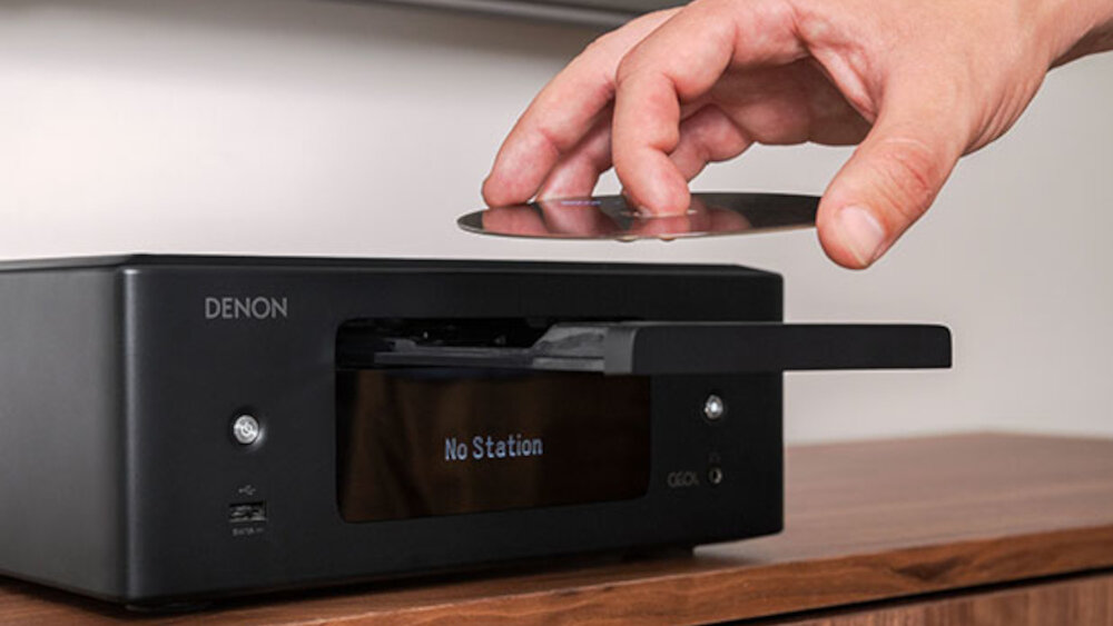 Czarny amplituner stereo z logo DENON, z wyświetlaczem pokazującym tekst \'No Station\'. Osoba wkłada płytę CD do urządzenia.