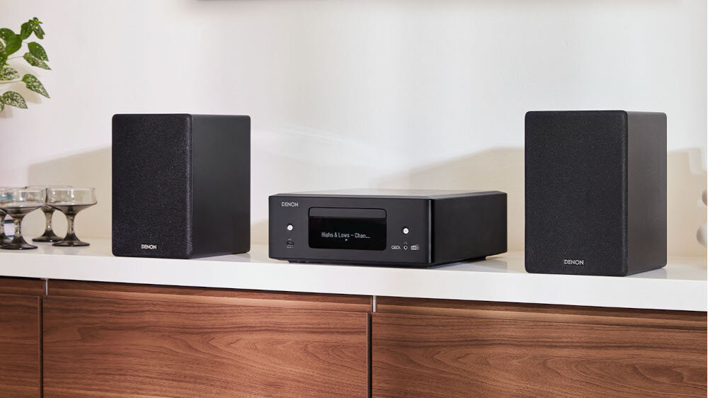 Na białej półce znajduje się czarny amplituner stereo marki DENON oraz dwa czarne głośniki. Obok amplitunera widoczne są trzy kieliszki na niskiej nóżce.