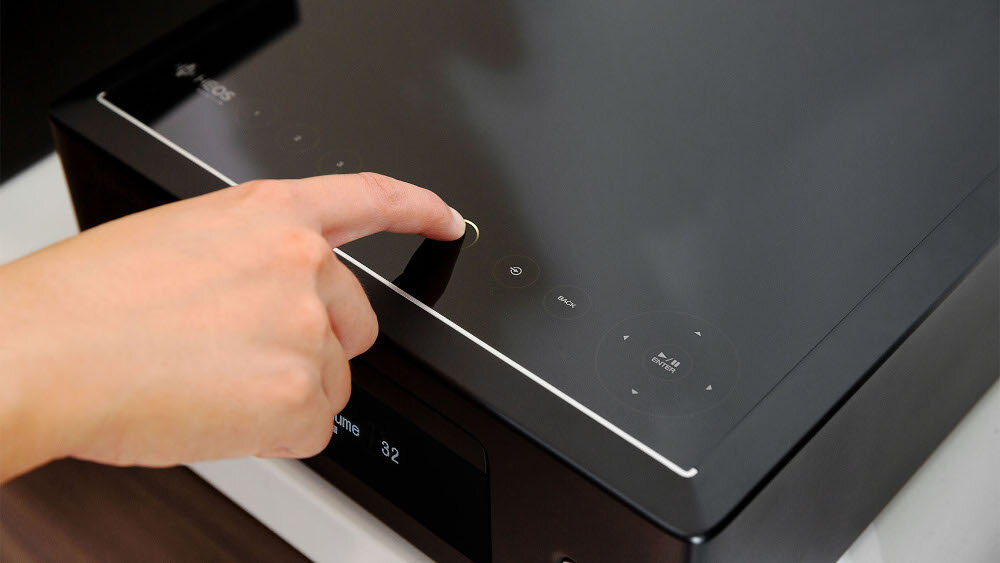 Osoba dotyka panelu dotykowego na czarnym amplitunerze stereo. Na wyświetlaczu widoczny jest napis \'Volume 32\'.