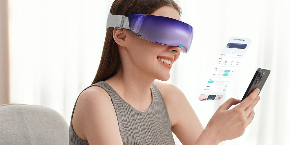 Osoba siedzi na krześle, nosząc fioletowy masażer do oczu i trzymając w ręku smartfon. Na ekranie telefonu widoczny jest interfejs aplikacji związanej z masażerem.