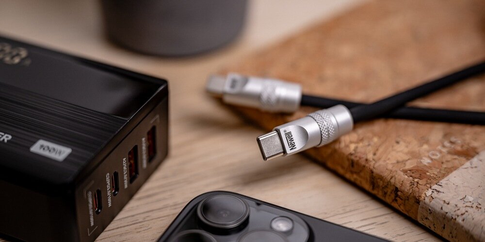 Fragment silikonowego kabla Newell na podkładce korkowej na drewnianym stole. Dookoła ładowarka, telefon i donica.