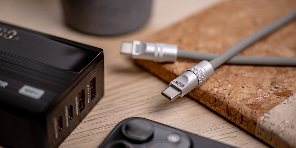 Fragment silikonowego kabla Newell na podkładce korkowej na drewnianym stole. Dookoła ładowarka, telefon i donica.