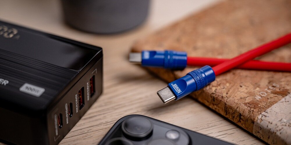 Fragment silikonowego kabla Newell na podkładce korkowej na drewnianym stole. Dookoła ładowarka, telefon i donica.