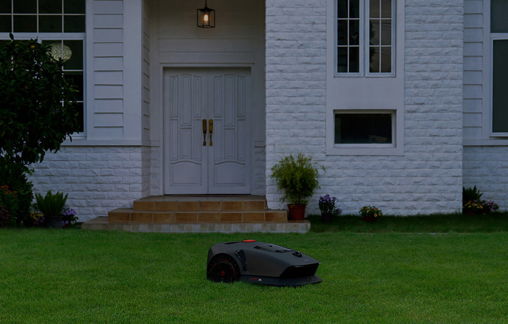 Robot koszący DREAME Mova 600 robot na trawie w tle biały dom wieczorna pora poziom hałasu nie przekracza 60 dB akumulator ekologiczne rozwiązanie akumulator o pojemności 2,5 Ah i napięciu 18 V czas pracy około 50 minut na jednym ładowaniu
