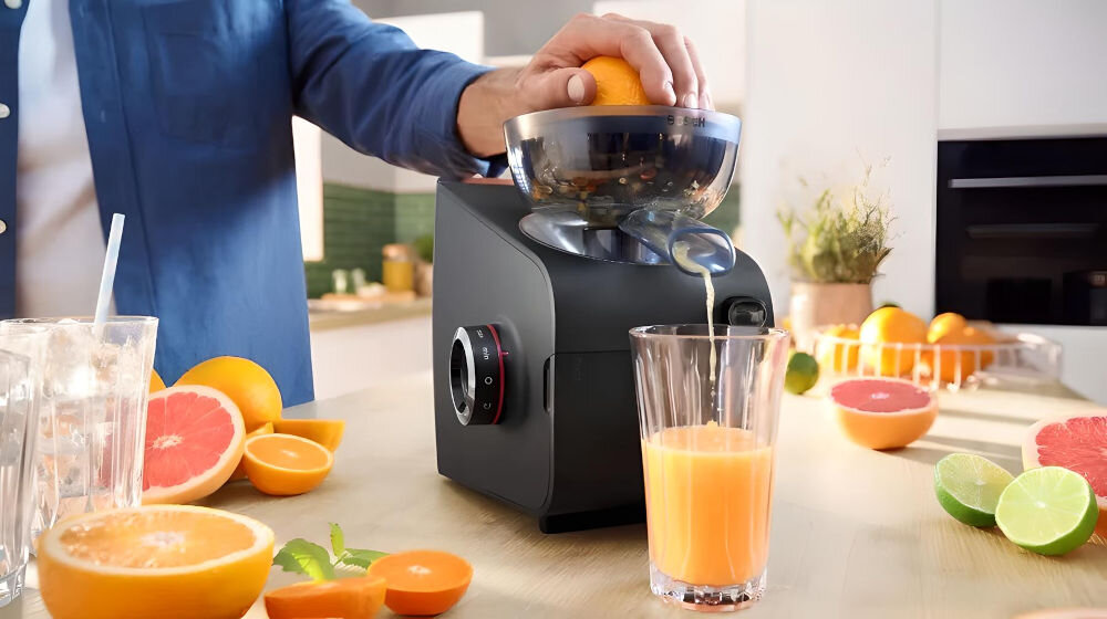 Maszynka do mięsa BOSCH MFWS682B maszynka pokrętło kuchenne tło ręką wkładające pomarańczę do wyciskarki pomarańczowy sok wlatujący do szklanki owoce cytrusowe leżące na blacie wielofunkcyjne urządzenie wyciskarka do cytrusów mielenie mięsa przygotowywanie świeżych soków łatwy montaż i demontaż mocny silnik maszynki szybkie efektywne wyciskanie soków pomarańcze cytryny grejpfruty praktyczny pomocnik w kuchni   