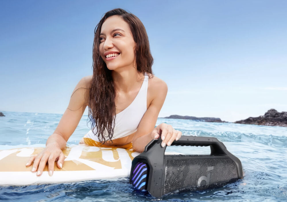 Głośnik mobilny SOUNDCORE Boom 2 Plus wodoodporny IPX7 unoszący się na wodzie, osoba trzymająca się deski surfingowej w wodzie , głosnik zamoczony w wodzie