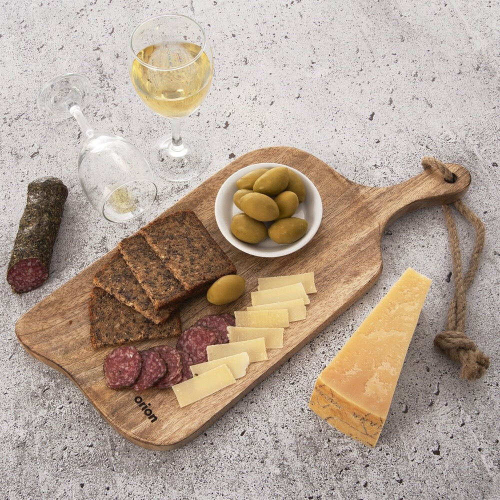 Na drewnianej desce do krojenia znajdują się plasterki chleba, salami, sera oraz miseczka z oliwkami. Obok deski stoi kieliszek z białym winem, a na blacie leży kawałek sera i butelka wina.