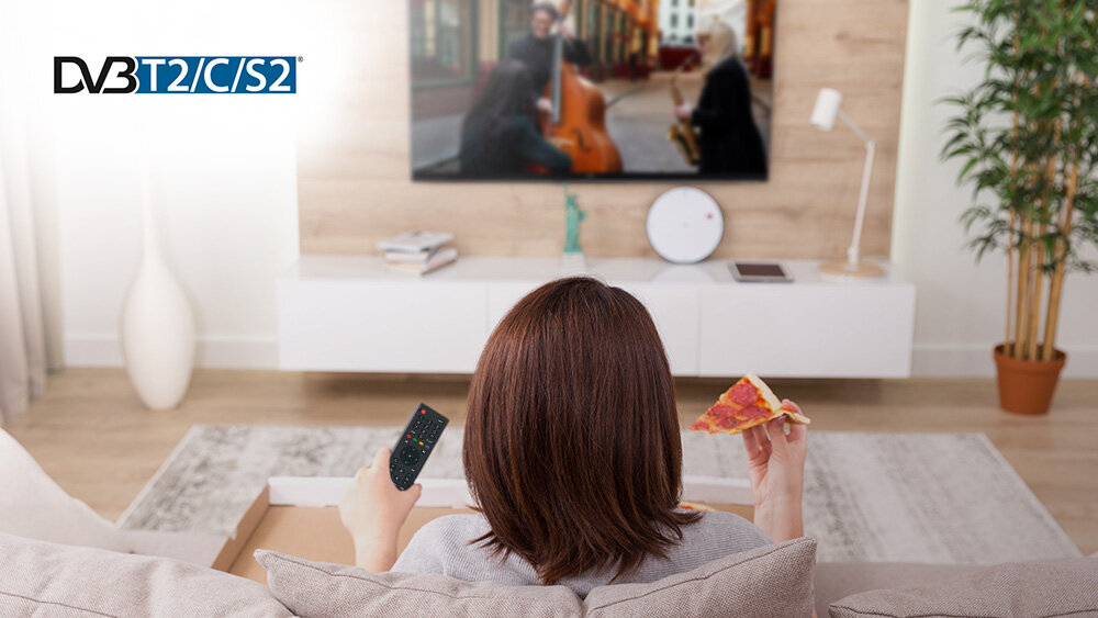 Telewizor JVC LT-32VDH5400 - Potrójny tuner DVB-T2/C/S2, kobieta trzymająca w ręku pilota i pizzę, telewizor powieszony na ścianie w salonie