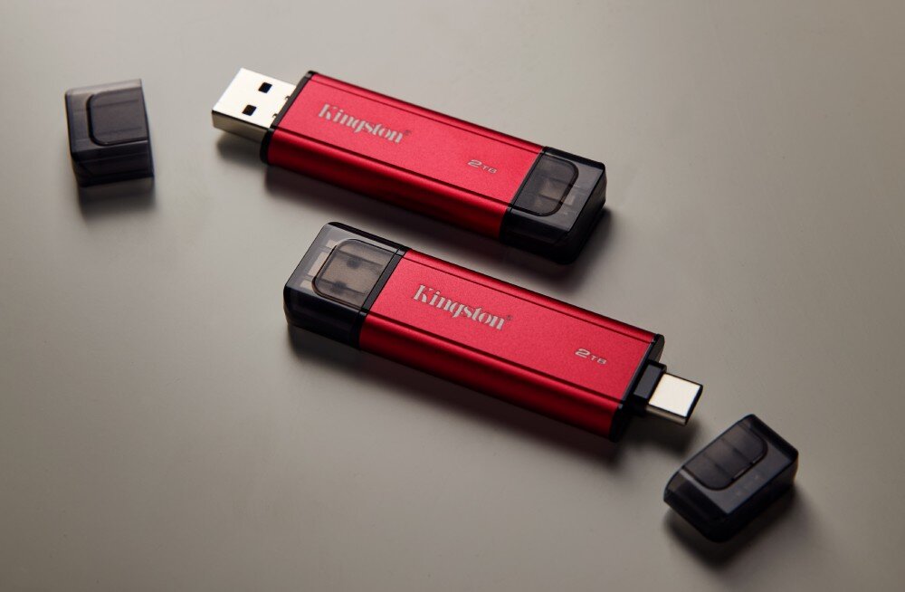 Dysk KINGSTON Dual Portable Dwa małe, podłużne urządzenia z napisami 'Kingston' i '2 TB', z których jedno ma odsłoniętą wtyczkę, leżą na białej powierzchni obok dwóch zatyczek, opis