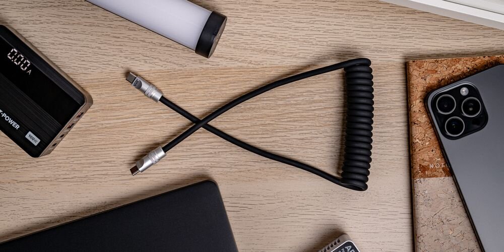Kabel Newell USB-C - USB-C 100 W, 1,5m, silikonowy, spiralny, na drewnianym blacie pośród sprzętów do ładowania.