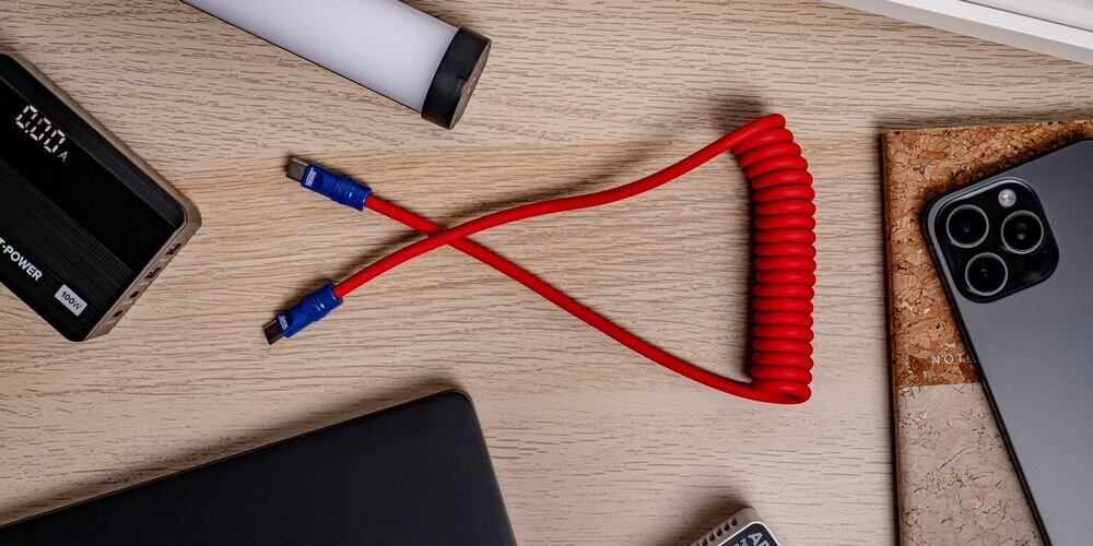 Kabel Newell USB-C - USB-C 100 W, 1,5m, silikonowy, spiralny, na drewnianym blacie pośród sprzętów do ładowania.
