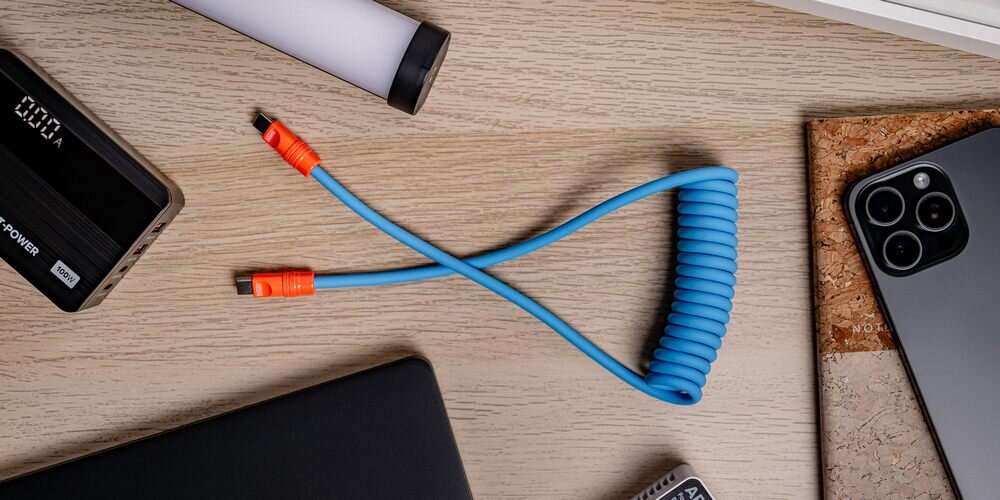 Kabel Newell USB-C - USB-C 100 W, 1,5m, silikonowy, spiralny, na drewnianym blacie pośród sprzętów do ładowania.