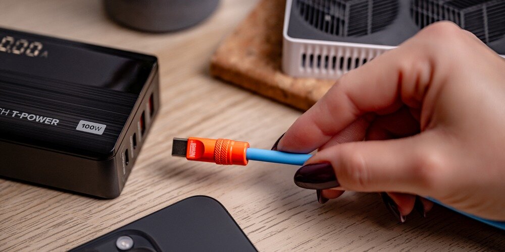 Kobieca dłoń zbliża końcówkę kabla Newell USB-C - USB-C 100 W, 1,5m do leżącej na drewnianym stole ładowarki.