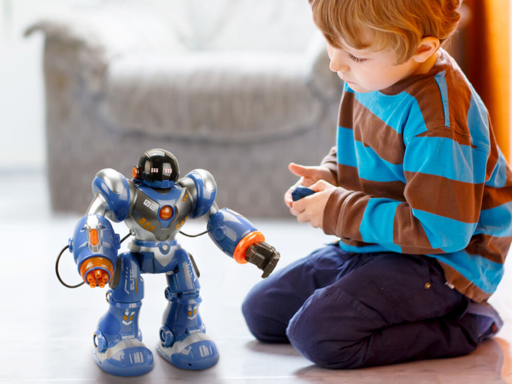 Na podłodze znajduje się niebiesko-srebrny robot z pomarańczowymi elementami, obok którego klęczy dziecko trzymające pilot. Robot ma czarną głowę z wyświetlaczem i jest w pozycji stojącej.