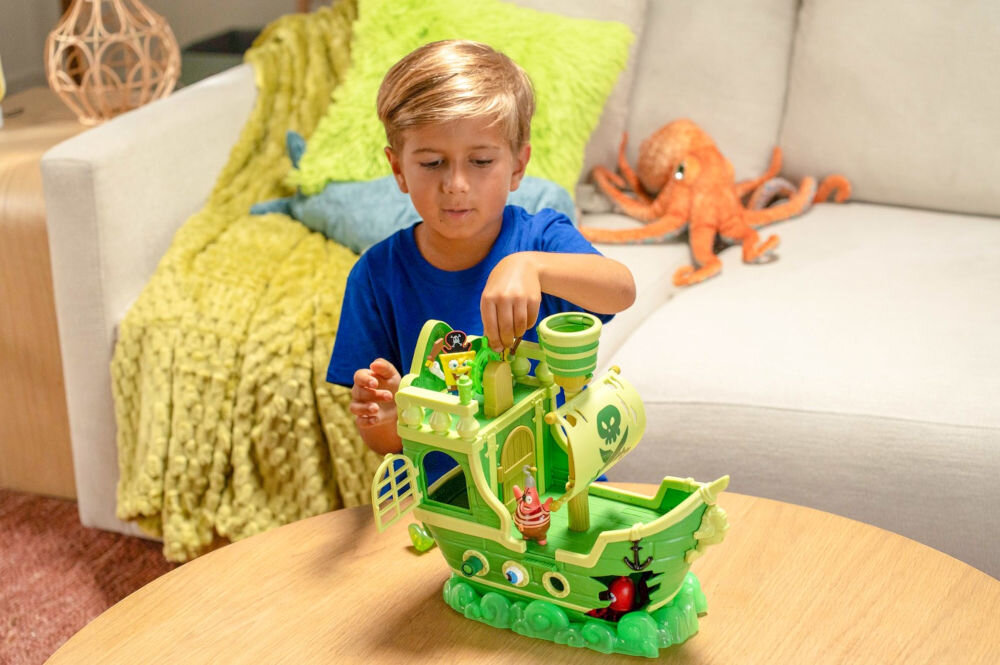 Zestaw figurek FUNRISE SpongeBob Latający Holender 73110 Chłopiec w niebieskiej koszulce bawiący się na drewnianym stole zabawkowym statkiem pirackim SpongeBob Kanciastoporty Latający Holender. Dziecko kręci zielonym kołem sterowym na mostku kapitańskim obok figurki SpongeBoba w stroju pirata. W tle widać jasną kanapę z pluszowymi zabawkami.
