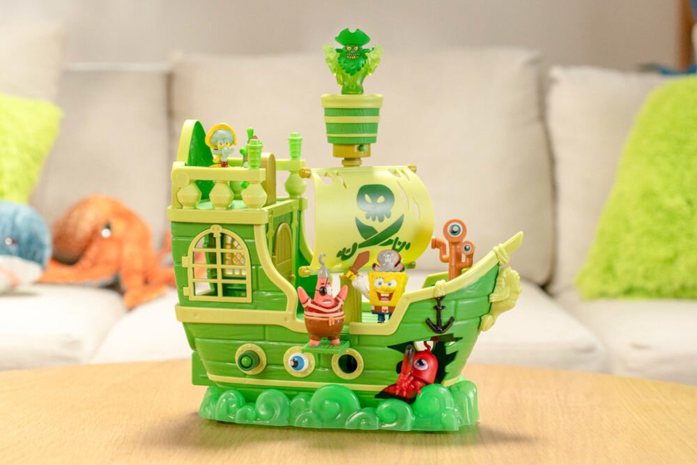 Zestaw figurek FUNRISE SpongeBob Latający Holender 73110 Widok z boku na zabawkowy statek piracki SpongeBoba pod nazwą Latający Holender. Pojazd jest zielono-żółty, ma żagiel z logo czaszki i skrzyżowanych kości oraz figurki piratów SpongeBoba i Patryka. Na szczycie masztu znajduje się świecąca, zielona figura ducha.